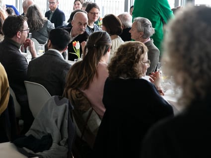 A large group of people sit at a long table chatting and eating a meal together.