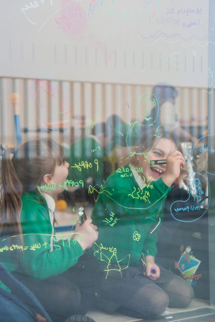 Two primary school children smile as the draw with coloured markers onto a glass window in the Foyle rooms.