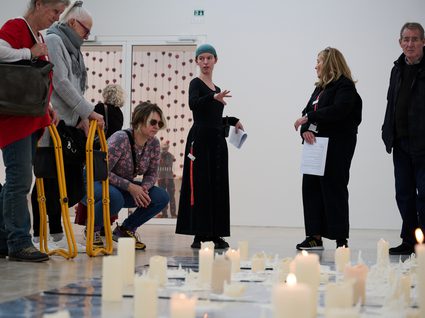 A group of people stand looking at an art installation made up of hundreds of burning candles sat on foil that covers the floor.