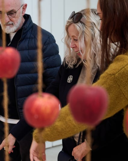 Three visitors stand behind a curtain of threaded apples.
