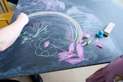 A child's hand draws across a chalk board with multicoloured images of flowers and a rainbow on.