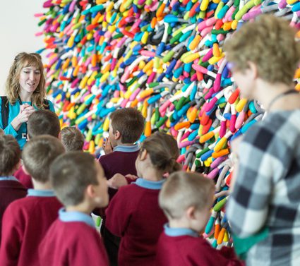 A group of children stand in front of a large, wall-based sculpture with soft multicoloured tentacles poking out from the wall